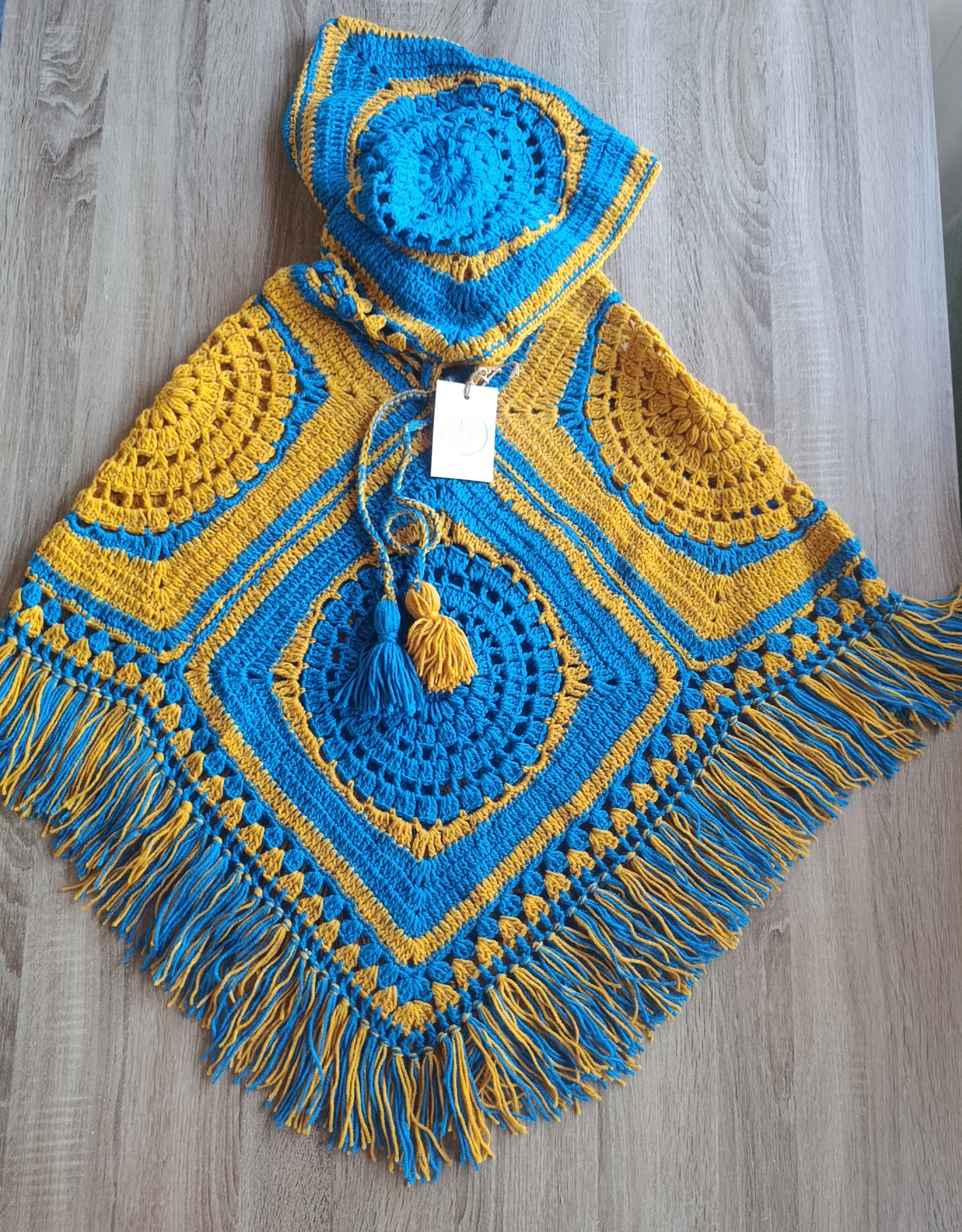 Poncho de crochê com capuz Kukini co. em azul e amarelo, destacando o tecido colorido e detalhes do design.